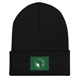 Puede incluir: Gorro negro con un parche verde que muestra la bandera de la Unión Africana. La bandera tiene un círculo dorado con un contorno blanco alrededor de una silueta blanca del continente africano.