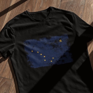 May include: A black t-shirt with a distressed blue and white print of the flag of Alaska. The flag features a single gold star in the upper left corner and eight gold stars in a constellation pattern in the lower right corner.