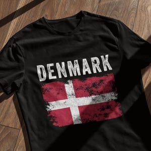 Könnte beinhalten: Schwarzes T-Shirt mit einem abgenutzten weißen Grafikdruck der dänischen Flagge und dem Wort "DENMARK" darüber.