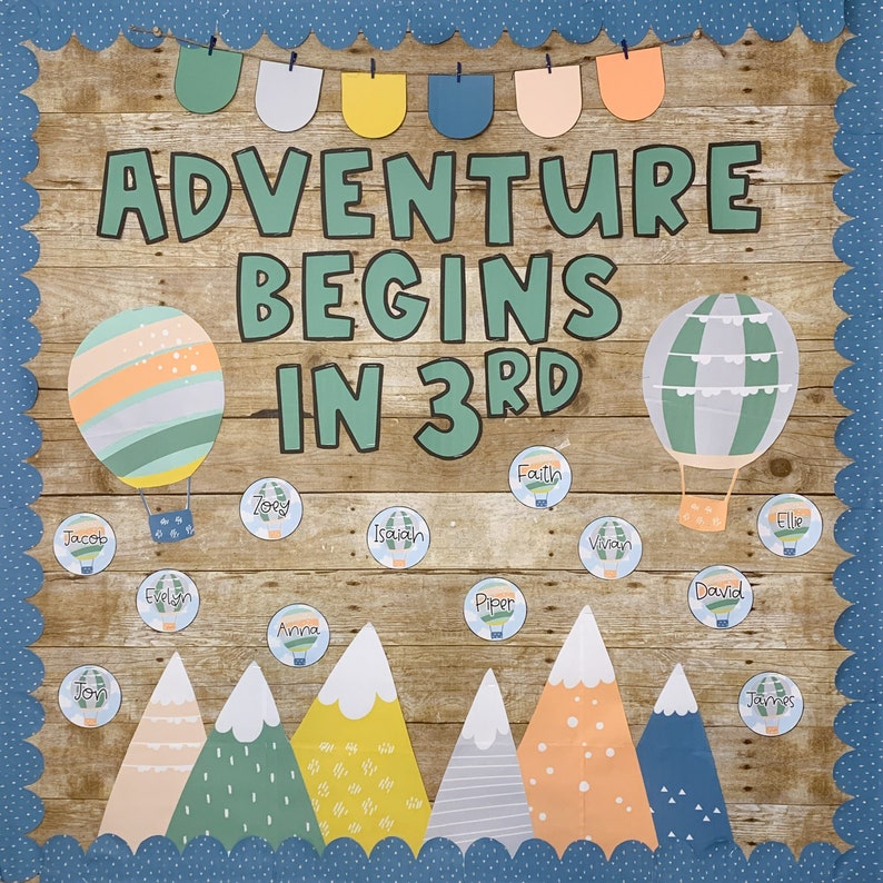 May include: A wooden bulletin board with the text "ADVENTURE BEGINS IN 3RD" in green letters. The board is decorated with colorful paper bunting, hot air balloons, and mountains. There are also small circles with names written on them.