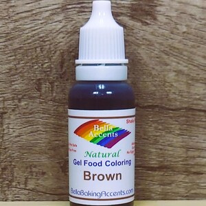 May include: A bottle of brown gel food coloring with a white cap and a label featuring a rainbow logo and the text "Bella Accents Natural Gel Food Coloring Brown". The bottle is against a wooden background.