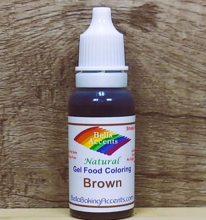 May include: A bottle of brown gel food coloring with a white cap and a label featuring a rainbow logo and the text "Bella Accents Natural Gel Food Coloring Brown". The bottle is against a wooden background.