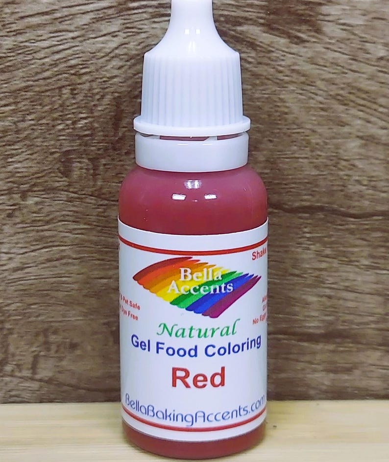May include: A bottle of red gel food coloring with a white cap and label. The label features a rainbow design and the words "Bella Accents Natural Gel Food Coloring Red". The bottle is against a wooden background.