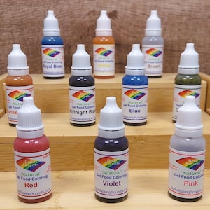 May include: A collection of natural gel food coloring bottles in various colors, including red, violet, and pink. Each bottle has a white cap and a label with the brand name and color. The bottles are arranged on wooden shelves.