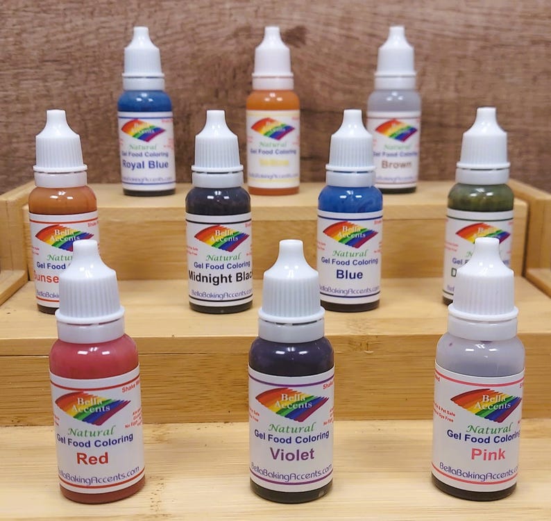 May include: A collection of natural gel food coloring bottles in various colors, including red, violet, and pink. Each bottle has a white cap and a label with the brand name and color. The bottles are arranged on wooden shelves.
