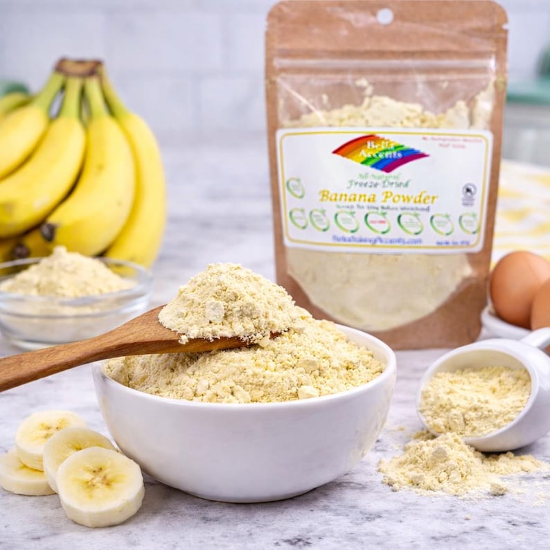 Banana powder in bowl with spoon on kitchen counter with pouch and fresh bananas for baking