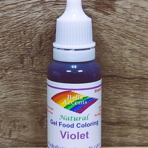 May include: A bottle of violet-colored natural gel food coloring. The bottle has a white cap and a label with the text "Bella Accents" and "Natural Gel Food Coloring Violet". The label also includes a rainbow design.