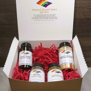 Puede incluir: Una caja de regalo con cuatro botellas de salsa de postre. La caja es blanca con un interior dorado y está llena de papel triturado rojo. Las botellas están etiquetadas como "Chocolate Awesome Sauce", "Sea Salt Caramel Awesome Sauce", "Caramel Awesome Sauce" y "White Chocolate Awesome Sauce".