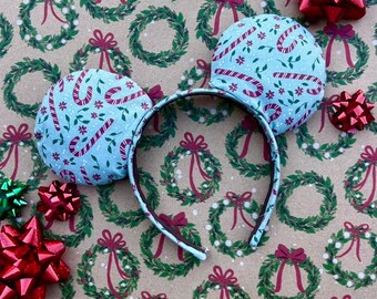 May include: A pair of blue and white mouse ears with a candy cane and floral pattern. The ears are on a brown background with a repeating pattern of green wreaths.
