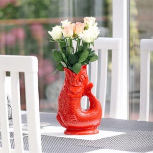 Könnte beinhalten: Eine leuchtend rote Keramikvase in Fischform, gefüllt mit weißen und pfirsichfarbenen Rosen. Die Vase hat ein detailliertes Schuppenmuster und einen geschwungenen Schwanzgriff. Die Blumen sind frisch und grün. Die Vase steht auf einem Tisch.