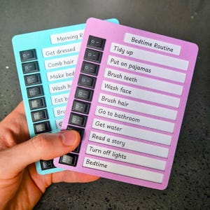 May include: Two colourful routine checklist boards are held in a hand. One is light blue and the other is pink. Each board has a series of toggle switches and labels for morning and bedtime routines. The text includes "Get dressed" and "Brush teeth."