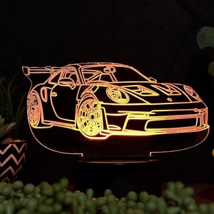 May include: A 3D printed acrylic lamp with a glowing orange outline of a sports car. The lamp is on a black base and is sitting on a table with a blue figurine and plants.