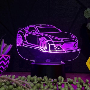 May include: A purple and black 3D printed night light shaped like a car. The car is a silhouette of a sports car with a large spoiler and wide tires. The night light is on a black base and is lit up with purple light.