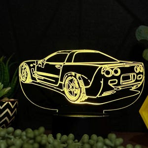 May include: A yellow 3D printed night light shaped like a Corvette sports car. The night light is on a black base and is illuminated from within.