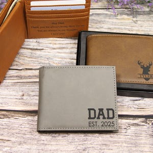 May include: Three leather wallets in shades of brown and gray are displayed on a wooden surface. One wallet is open, revealing card slots and text. Another is in a black box, featuring a deer graphic and a name. The third wallet has "DAD EST. 2025" printed on it.