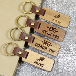 Puede incluir: Cuatro llaveros de madera con correas de cuero. Los llaveros presentan texto y símbolos grabados, incluyendo "Zachary", "Kyle #20", "Coach Tim" y "Valerie".