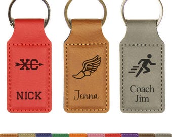 May include: Three leather keychains in red, tan, and gray. Each keychain has a metal ring and a rectangular tag with stitched edges. The red keychain says "XC" and "NICK". The tan keychain has a winged shoe and "Jenna". The gray keychain says "Coach Jim".