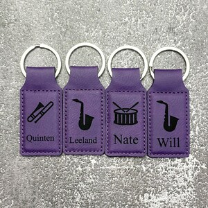 May include: Four purple keychains with silver rings. Each keychain has a different musical instrument icon: a trombone, a saxophone, a drum set, and a saxophone. Each keychain has a name: Quinten, Leeland, Nate, and Will.