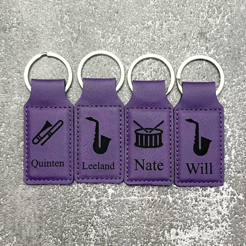 May include: Four purple keychains with silver rings. Each keychain has a different musical instrument icon: a trombone, a saxophone, a drum set, and a saxophone. Each keychain has a name: Quinten, Leeland, Nate, and Will.