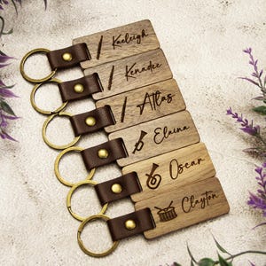Puede incluir: Llaveros de madera personalizados con nombres y dise&ntilde;os de instrumentos musicales. Cada llavero tiene una correa de cuero marr&oacute;n y un anillo dorado. Nombres: Kaeligh, Kennadee, Atlas, Elaina, Oscar y Clayton.