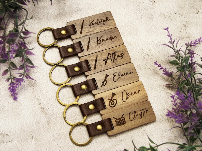 Puede incluir: Llaveros de madera personalizados con nombres y dise&ntilde;os de instrumentos musicales. Cada llavero tiene una correa de cuero marr&oacute;n y un anillo dorado. Nombres: Kaeligh, Kennadee, Atlas, Elaina, Oscar y Clayton.