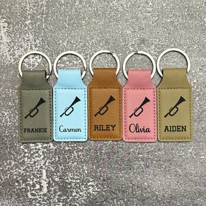 May include: Five personalized keychains in various colors: gray, light blue, brown, pink, and olive green. Each keychain features a black trumpet graphic and a name: Frankie, Carmen, Riley, Olivia, and Aiden. The keychains have silver key rings.
