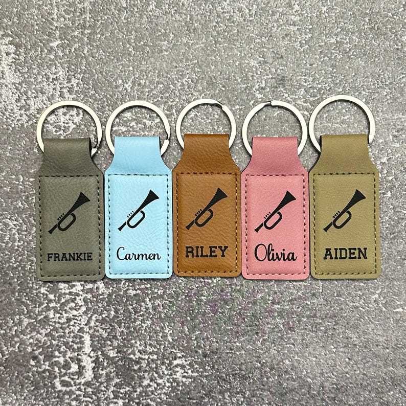 May include: Five personalized keychains in various colors: gray, light blue, brown, pink, and olive green. Each keychain features a black trumpet graphic and a name: Frankie, Carmen, Riley, Olivia, and Aiden. The keychains have silver key rings.