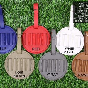 May include: A collection of round luggage tags in various colors: blue, red, white marble, light brown, gray, and rawhide. Each tag has a strap and a section with five vertical slots. The tags are displayed on a green grassy surface.
