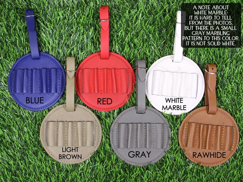 May include: A collection of round luggage tags in various colors: blue, red, white marble, light brown, gray, and rawhide. Each tag has a strap and a section with five vertical slots. The tags are displayed on a green grassy surface.