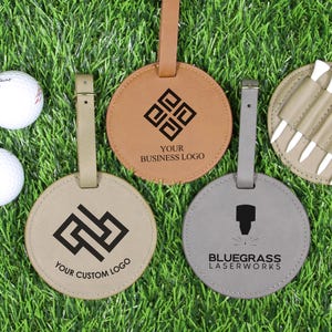 May include: Golf-themed accessories on green grass. Includes three white golf balls, and four leather items: three round luggage tags with custom logos and a holder with golf tees. The tags are tan, brown, and gray.