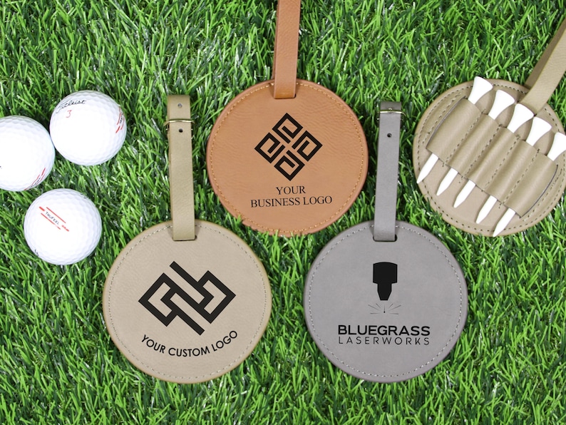 May include: Golf-themed accessories on green grass. Includes three white golf balls, and four leather items: three round luggage tags with custom logos and a holder with golf tees. The tags are tan, brown, and gray.