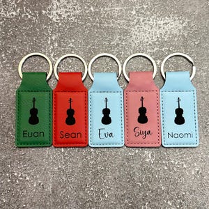 May include: Five colorful keychains with silver rings. Each keychain has a rectangular tag with a black violin silhouette and a name. The tags are green, red, light blue, pink, and blue.