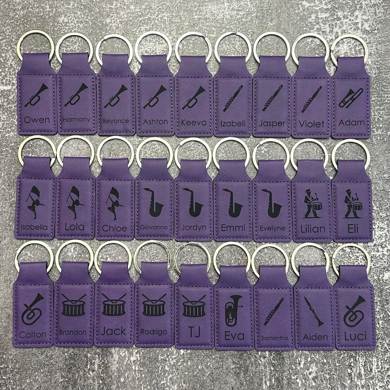 May include: A collection of purple keychains with silver rings. Each keychain features a black musical instrument graphic and a name. Instruments include trumpets, flutes, saxophones, drums, and a tuba. Names include Owen, Harmony, Beyonce, and others.