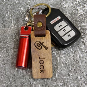 Puede incluir: Un conjunto de llaves y llaveros. Incluye un mando de coche, un tubo de metal rojo y un llavero de madera grabado con un instrumento musical y el nombre "Jack". El llavero de madera est&aacute; sujeto a una correa de cuero marr&oacute;n.