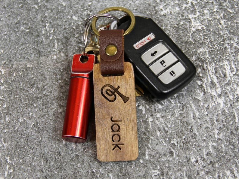 Puede incluir: Un conjunto de llaves y llaveros. Incluye un mando de coche, un tubo de metal rojo y un llavero de madera grabado con un instrumento musical y el nombre "Jack". El llavero de madera est&aacute; sujeto a una correa de cuero marr&oacute;n.