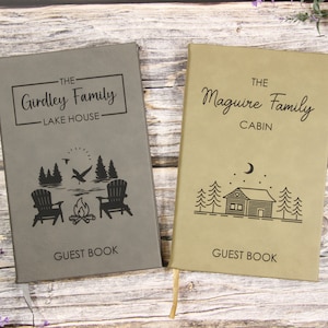 May include: Two gray and brown leather guest books with personalized text. The first book says "The Girdley Family Lake House" with a campfire and two chairs. The second book says "The Maguire Family Cabin" with a cabin and trees.