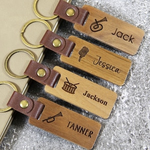 May include: Four wooden keychains with brown leather straps and brass rings. Each keychain is engraved with a name and a musical instrument: Jack with a trumpet, Jessica with a microphone, Jackson with a drum, and Tanner with a trumpet.
