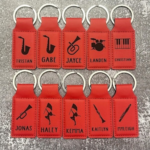 May include: A collection of ten red keychains with silver key rings. Each keychain features a black musical instrument graphic and a name. Instruments include a saxophone, trumpet, drums, piano keys, and a flag twirler.