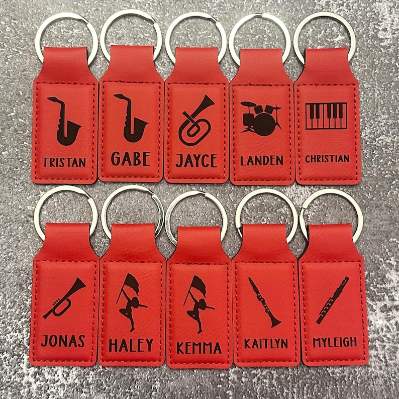 May include: A collection of ten red keychains with silver key rings. Each keychain features a black musical instrument graphic and a name. Instruments include a saxophone, trumpet, drums, piano keys, and a flag twirler.