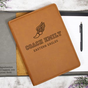 May include: A tan leather portfolio with the text "COACH EMILY EASTERN EAGLES" and a winged shoe logo. The portfolio is open, revealing a notepad and pen. The text "Opportunities don't happen, you create them" is on the left side.