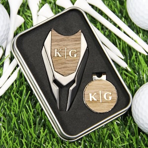 May include: A personalized golf tool set with a divot repair tool and ball marker, both featuring the initials "KG" in a wooden inlay. The set is presented in a metal tin, alongside golf balls and tees on a bed of green grass.