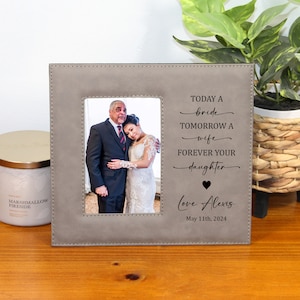 May include: A gray faux leather picture frame with a black engraved message that reads "Today a bride, tomorrow a wife, forever your daughter. Love, Alexis, May 11th, 2024". The frame has a photo of a bride and groom.