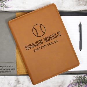 May include: A tan leather portfolio with a baseball graphic and the text "COACH EMILY EASTERN EAGLES." The portfolio is open, revealing a notepad and pen. The portfolio is on a gray and tan background with the text "Opportunities don't happen, you create them."