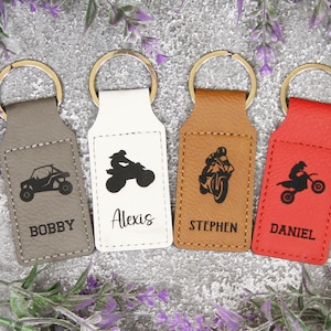 May include: Four leather keychains with different colored leather and personalized names. Each keychain features a different vehicle design: an ATV, a motorcycle, a dirt bike, and a race car. The names on the keychains are Bobby, Alexis, Stephen, and Daniel.