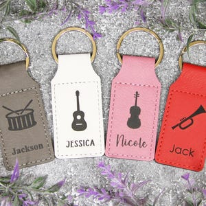 May include: Four keychains with gold rings. The keychains are in gray, white, pink, and red. Each keychain has a musical instrument graphic: a drum, guitar, violin, and trumpet. Each keychain has a name: Jackson, Jessica, Nicole, and Jack.