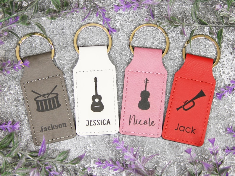 May include: Four keychains with gold rings. The keychains are in gray, white, pink, and red. Each keychain has a musical instrument graphic: a drum, guitar, violin, and trumpet. Each keychain has a name: Jackson, Jessica, Nicole, and Jack.
