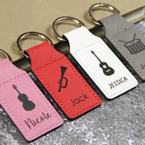 May include: Four personalized keychains in pink, red, white, and gray. Each keychain features a musical instrument silhouette and a name: violin/Nicole, trumpet/Jack, guitar/Jessica, and drums/Jackson. Keyrings are silver.