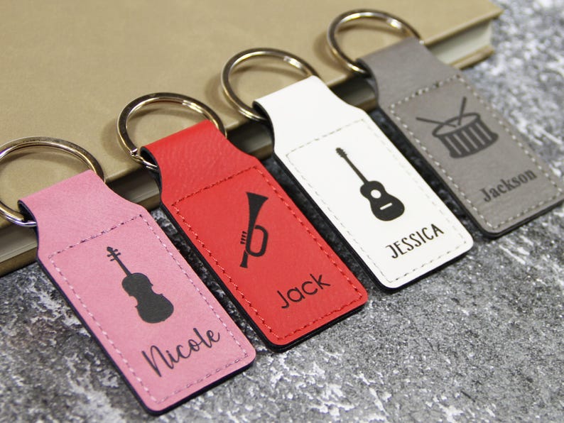 May include: Four personalized keychains in pink, red, white, and gray. Each keychain features a musical instrument silhouette and a name: violin/Nicole, trumpet/Jack, guitar/Jessica, and drums/Jackson. Keyrings are silver.