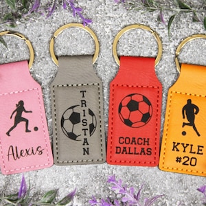 May include: Four personalized soccer keychains in pink, gray, red, and orange. Each keychain features a gold ring and a rectangular tag with a soccer-themed design and a name or text. The keychains are decorated with a soccer player silhouette, soccer ball, and text.