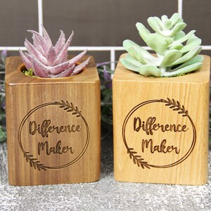 May include: Two wooden planter boxes with the text "Difference Maker" engraved on the front. One planter box has a pink succulent and the other has a green succulent.